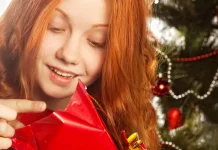 52 Best Tween Christmas Gifts for 2026, Curated By Experts Teen girl happily opening a red-wrapped Christmas present by the tree